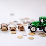 Miniatura de trator verde ao lado de pilhas de moedas sobre uma superfície clara.