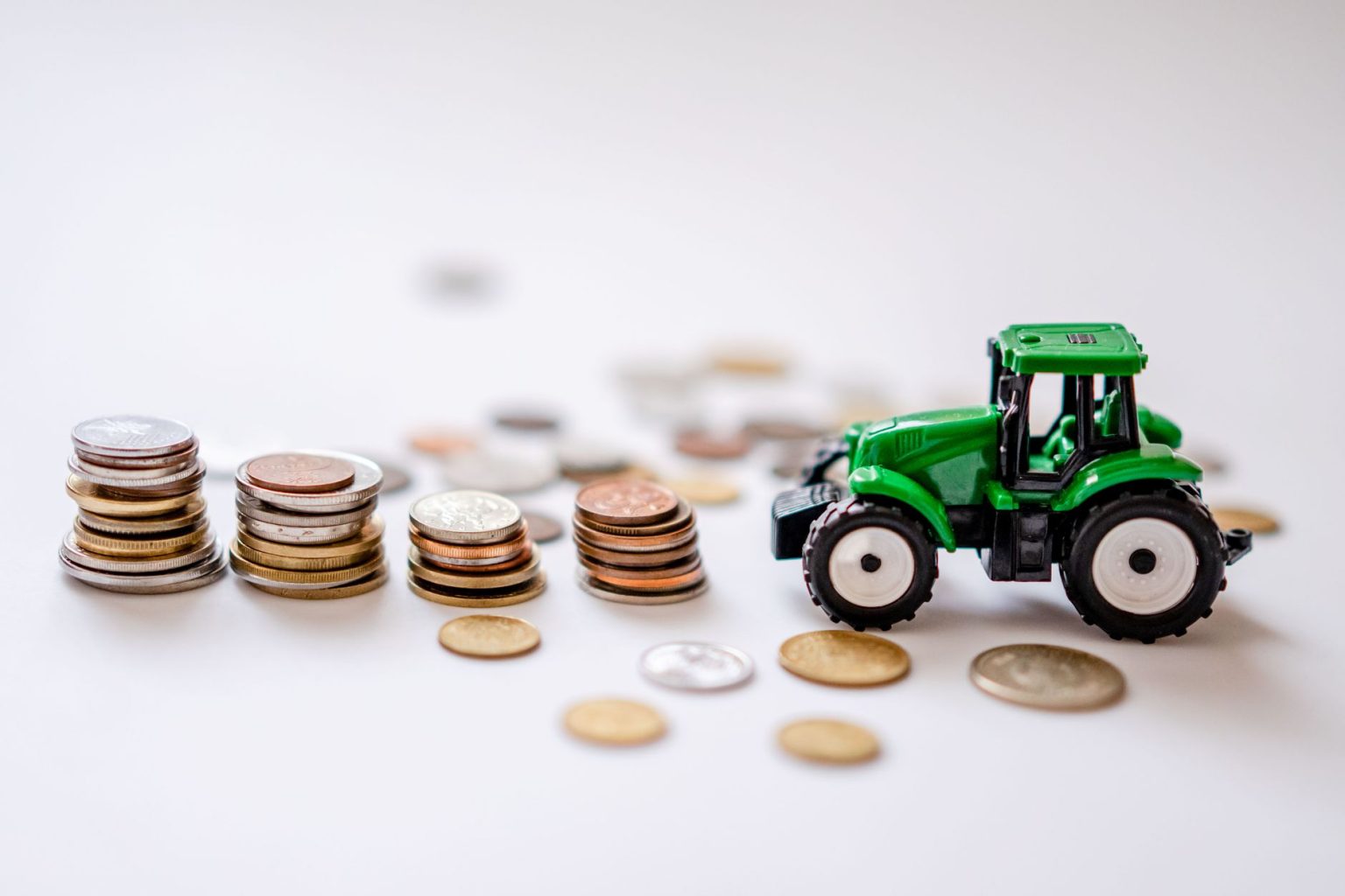 Miniatura de trator verde ao lado de pilhas de moedas sobre uma superfície clara.