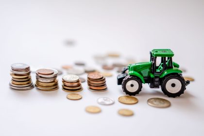 Miniatura de trator verde ao lado de pilhas de moedas sobre uma superfície clara.