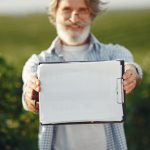 Agricultor idoso, usando óculos, segura uma prancheta em meio a uma plantação verde.