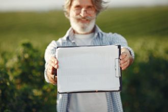 Agricultor idoso, usando óculos, segura uma prancheta em meio a uma plantação verde.