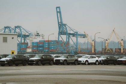 Fileira de automóveis estacionados em um terminal portuário, com guindastes e contêineres ao fundo