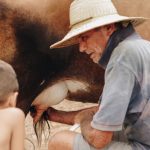 Homem idoso e criança ordenham uma vaca ao ar livre em uma propriedade rural no Brasil.