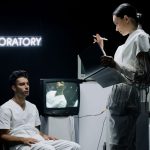 Dois pesquisadores analisam dados em um laboratório futurista enquanto conduzem um estudo com um participante.