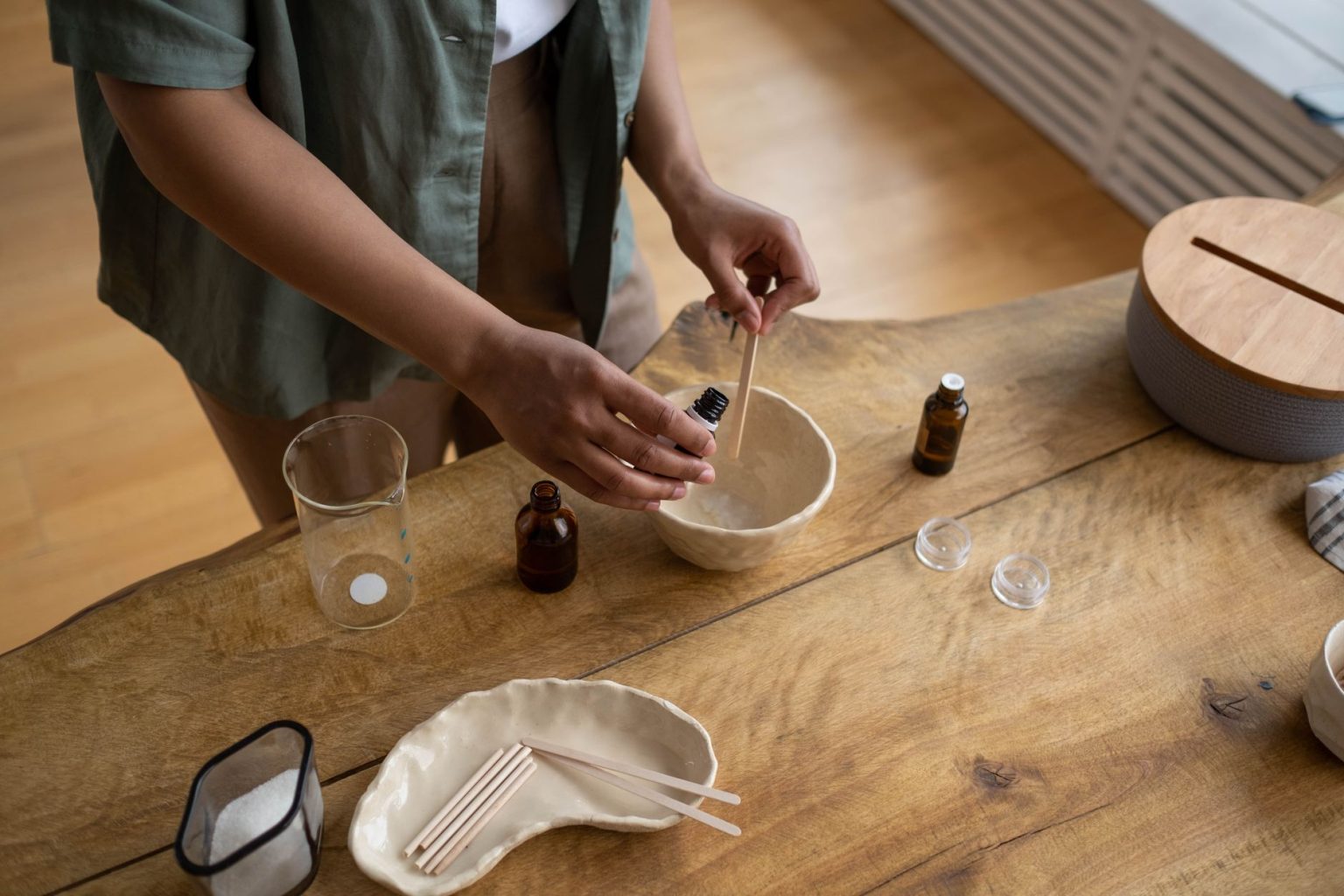 Mãos preparando artesanalmente um bálsamo labial em um recipiente, em ambiente interno.