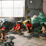 Quatro mulheres trabalham juntas na confecção de peças em uma oficina, cercadas por materiais e ferramentas.