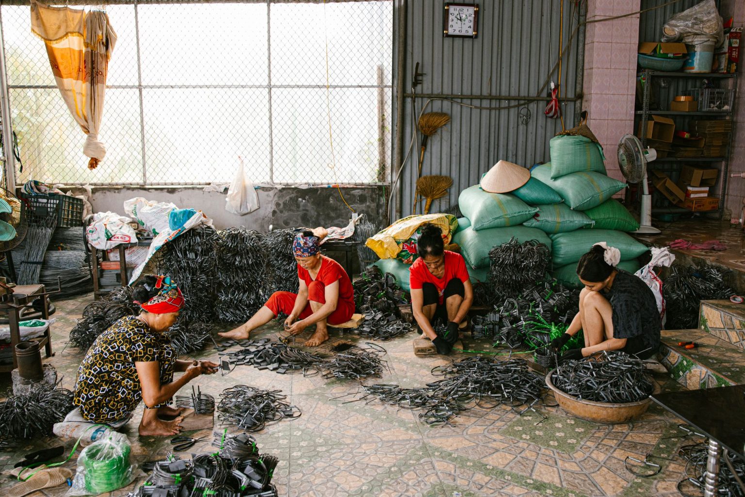 Quatro mulheres trabalham juntas na confecção de peças em uma oficina, cercadas por materiais e ferramentas.